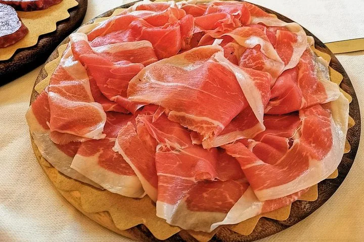 A round wooden board piled with thin, folded slices of pink Prosciutto di Parma